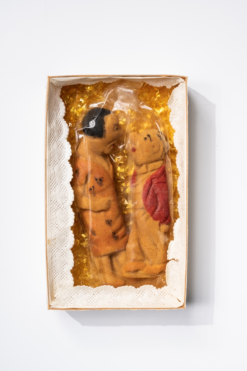 Konfektyr av marsipan eller tjockare deg formad till en kvinnofigur och en mansfigur (Selma och Lilla Fridolf). Kvinnofiguren har orangefärgad klänning med svart mönster. Mansfiguren har rödfärgad jacka. Figurerna ligger i påse av spröd plast (sprucken), på bädd av gula stanniolremsor i pappask (låda med lock). Runt lådans kanter är en spetsremsa av papper fastsatt.