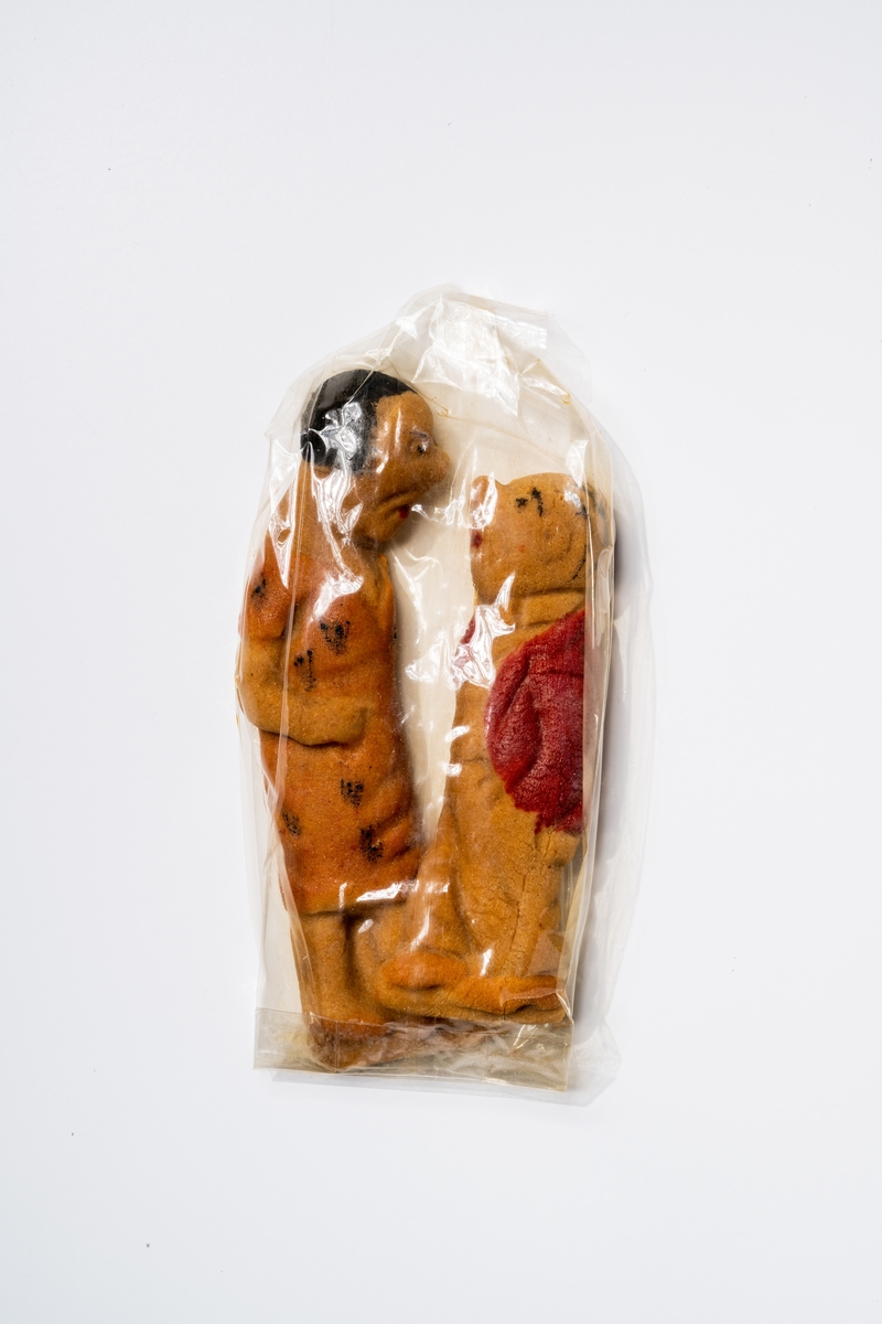 Konfektyr av marsipan eller tjockare deg formad till en kvinnofigur och en mansfigur (Selma och Lilla Fridolf). Kvinnofiguren har orangefärgad klänning med svart mönster. Mansfiguren har rödfärgad jacka. Figurerna ligger i påse av spröd plast (sprucken), på bädd av gula stanniolremsor i pappask (låda med lock). Runt lådans kanter är en spetsremsa av papper fastsatt.
