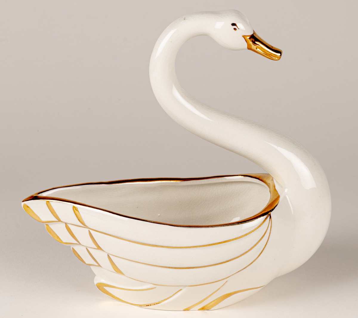 Prydnadsfigur eller skål i form av en svan, flintgods, modell A, dekorserie Aladdin; vit glasyr med handmålat guld. Aladdin producerades under åren 1941-1943. Oklart vem formgivaren är.