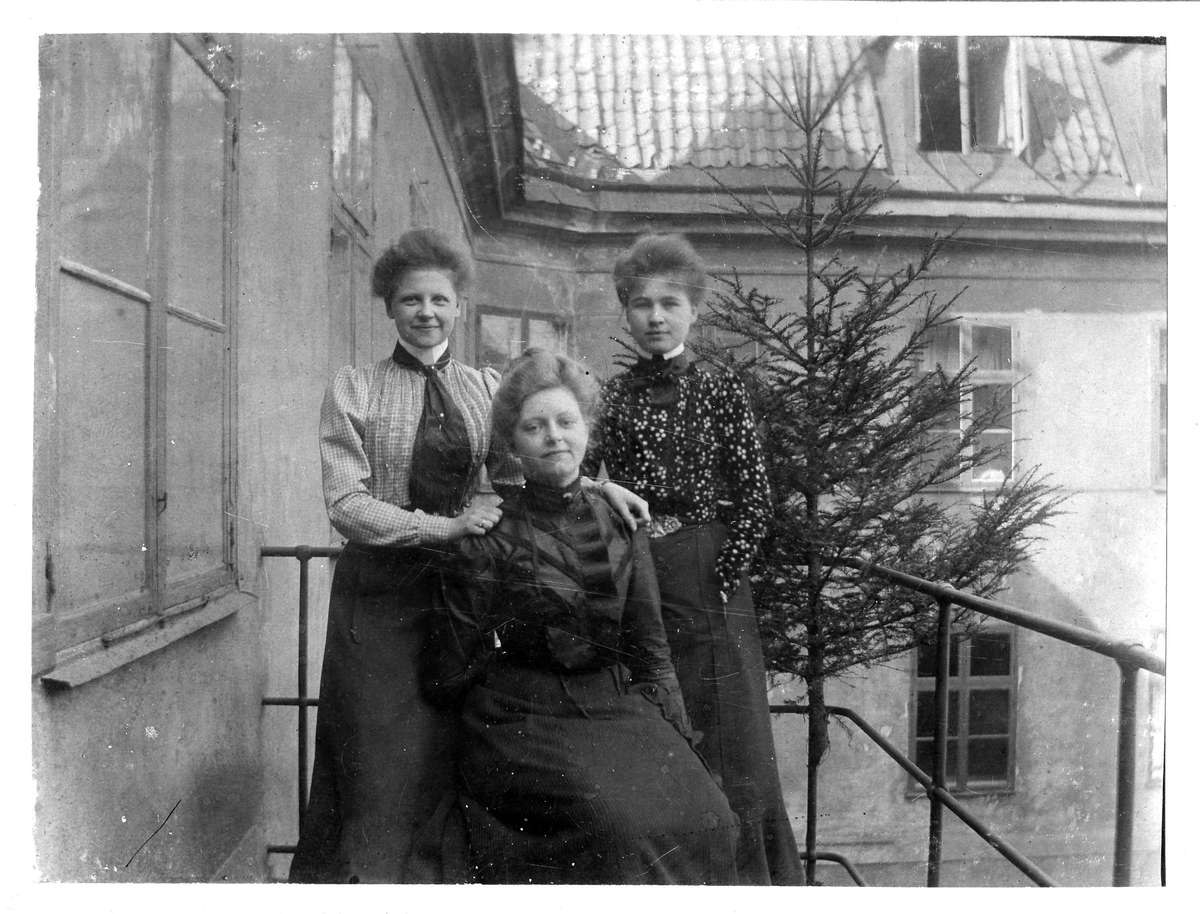 Bib Adelsköld och två okända kvinnor fotograferade på en balkong in mot vad som ser ut att vara en innergård. I bakgrunden står en gran.