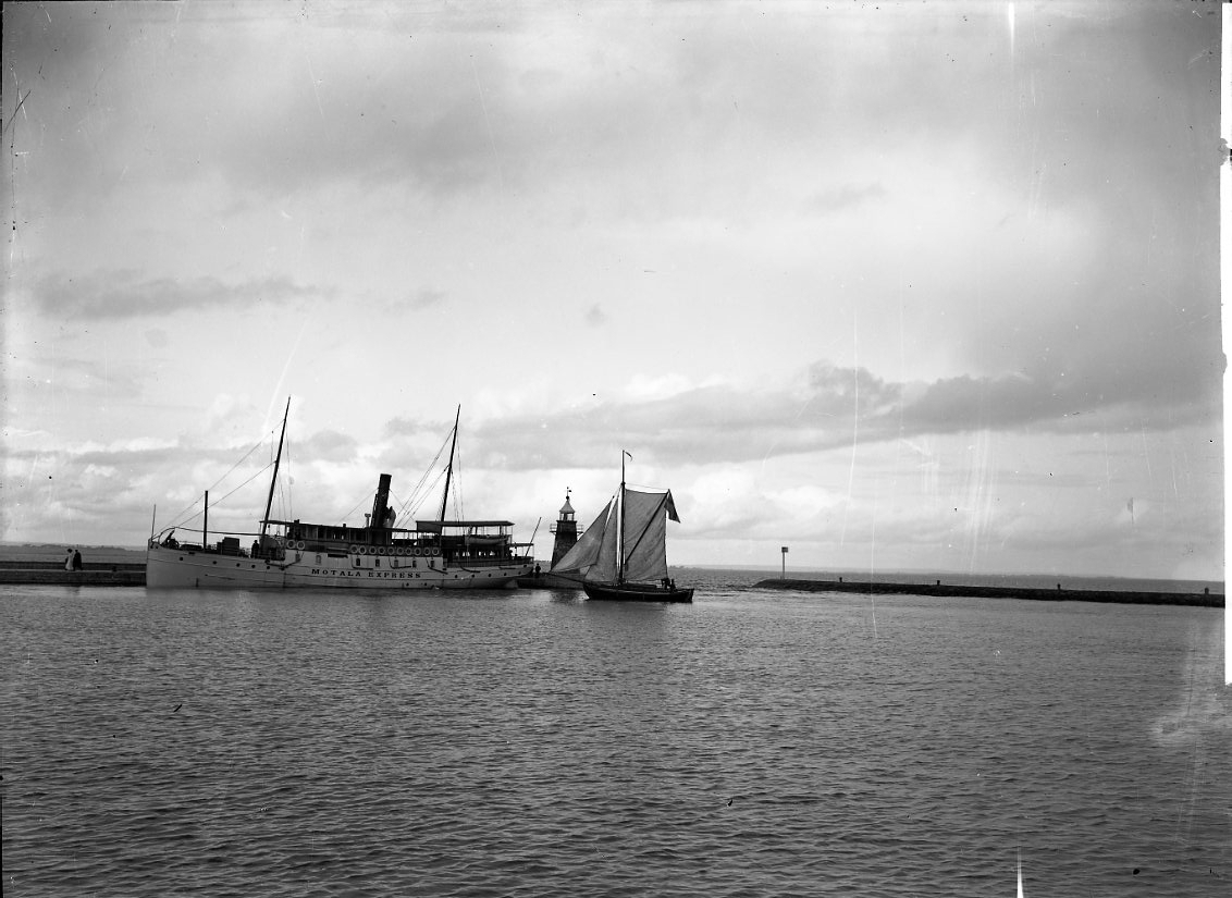 Ångbåten Motala Express ligger vid kaj i Gränna hamn. En segelbåt med satta segel till höger.