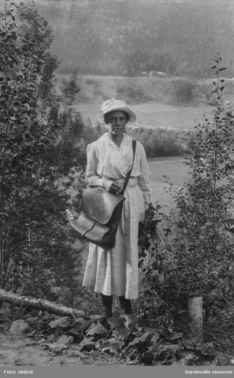 Porträtt av postiljon Hulda Höglund (senare gift Näsman) i Järkvissle, som började sin tjänst som lantbrevbärare 1917. Sträckan var Järkvissle-Västanå-Korsåmon och hon hämtade posten hos Larssons i Järkvissle för att sedan antingen gå eller cykla de tre milen tur och retur, på vintern nyttjades spark. Färjan i Järkvissle underlättade arbetet, tidigare var det till att ro över älven. I bakgrunden på södra sidan Indalsälven syns gården på Högland som var Huldas hem. Ur en samling som tillhört Sellén, Järkvissle.