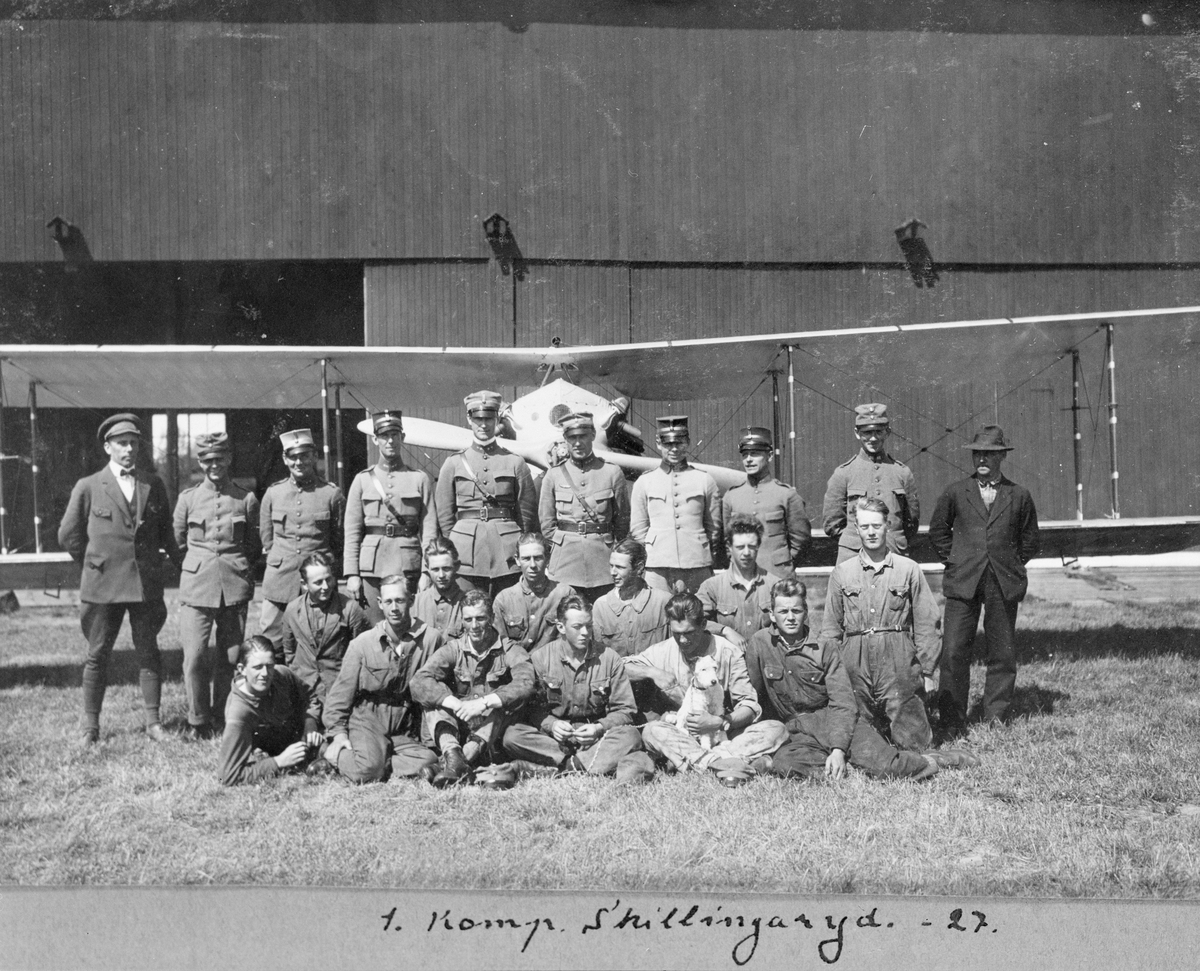 Gruppbild av militärer tillhörande 1. kompaniet Skillingaryd, 1927. 22 officerare och flygtekniker samt en hund framför flygplan J 2. I bakgrunden en hangar.