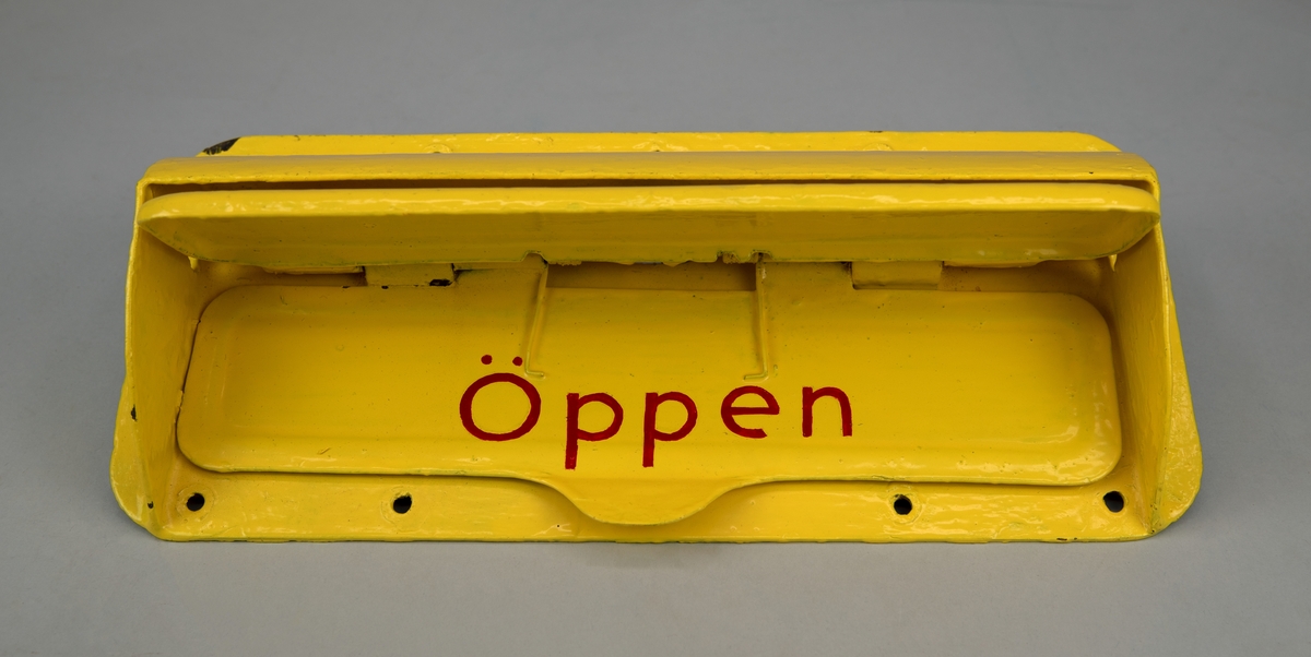 Rektangulärt brevinkast av gulmålad metall. Mitt på brevinkastet finns en öppning för brev. Två luckor, på den ena står det "Öppen", på den andra står det "Stängd" målat i rött. 10 st hål för fastsättning.