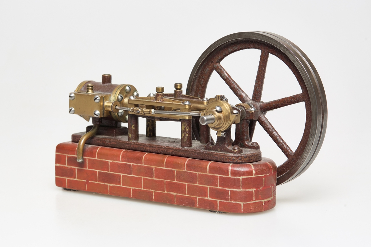 Modell av dampmaskin. Maskin av jern og messing, står på en tre base. Treverket er malt så det ser ut som murstein. Motoren driver ett hjul. En syllinder, stasjonær.