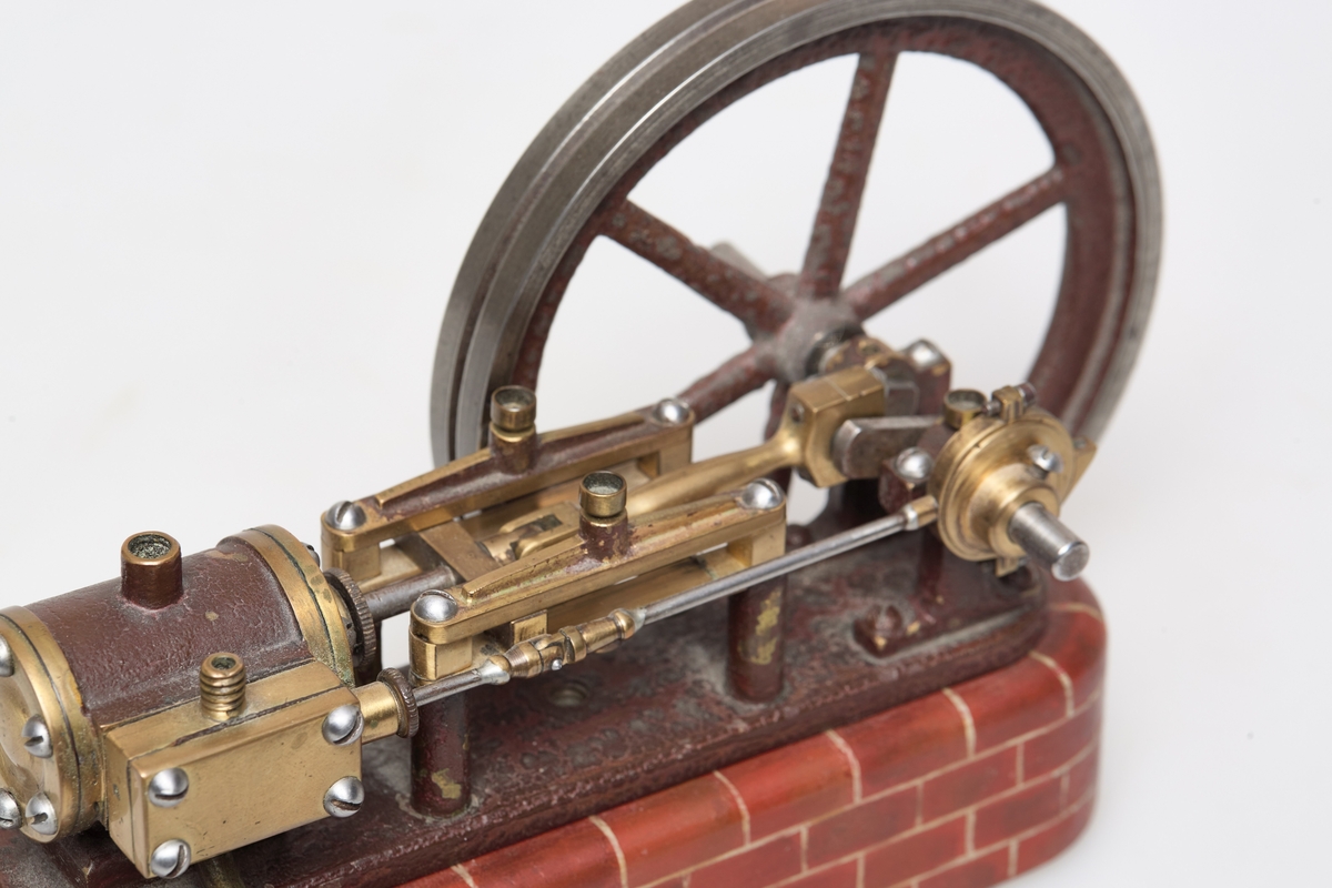 Modell av dampmaskin. Maskin av jern og messing, står på en tre base. Treverket er malt så det ser ut som murstein. Motoren driver ett hjul. En syllinder, stasjonær.