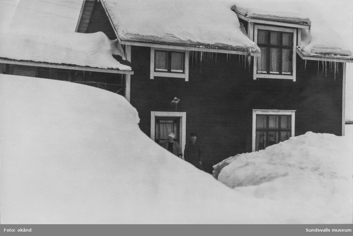 Framför dörren till ett trähus står ett äldre par. Huset omges av enorma snödrivor och det hänger långa istappar från taket. På bildens baksida står: Nybo Ortviken. Detta är fotograferat d. 2/2 26. Ur en samling från Skönsberg.