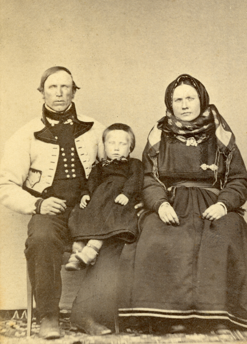 Familiegruppe i enkelt fotoatelier:
Kittil Olsson Bakås og Astrid Halvorsdotter Skogen g. Bakås, med sonen Halvor Kittilsson Bergland.  Tatt 1868/69