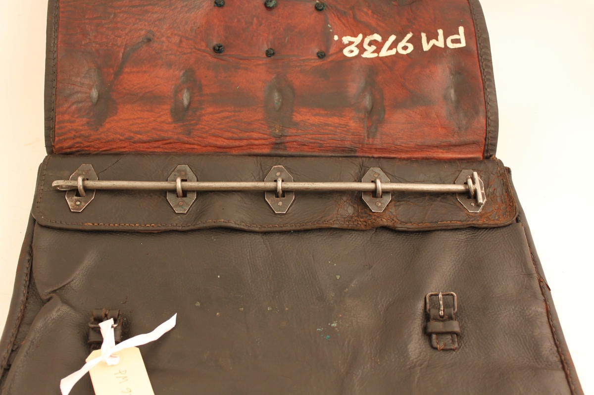 Väska av svärtat läder med klaff och två remmar och hålspännen. Låses med järnten (saknas). På klaffen en mässingsbricka märkt med "No 700".
