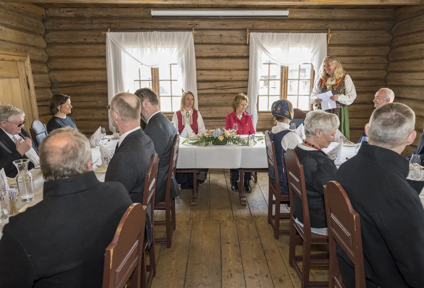 En samling personer sitter i en hesteskoformasjon i et rom med tømmervegger, alle er pyntet i dress eller bunad, en person står oppreist og holder et papir ved brystet