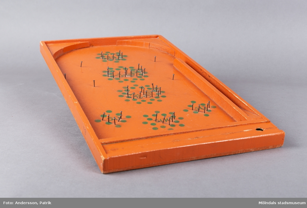 Fortunaspel bestående av en bottenplatta av bemålad masonit med kanter och ett fack av bemålat trä.
I facket finns en kort kö, två fotpinnar och metallkulor. Planen är försedd med spikar och en grön dekor som anger värdet. På vänster ytterkant är initialerna E.L skrivna med blyerts.
Spelet är handgjort och målat med orange färg. Punkter och siffror är målade i grön färg. Längst ned på spelet fins ett litet fack med speltillbehör. I facket finns en kort kö, två fotpinnar och metallkulor. Facket har ett lock som kan skjutas in. Spelet är förmodligen tillverkat på 50-talet och är en föregångare till flipperspelet. Poäng uppnås genom att kulor skjuts iväg med en pinne  och därefter hamnar i fack med olika värden.