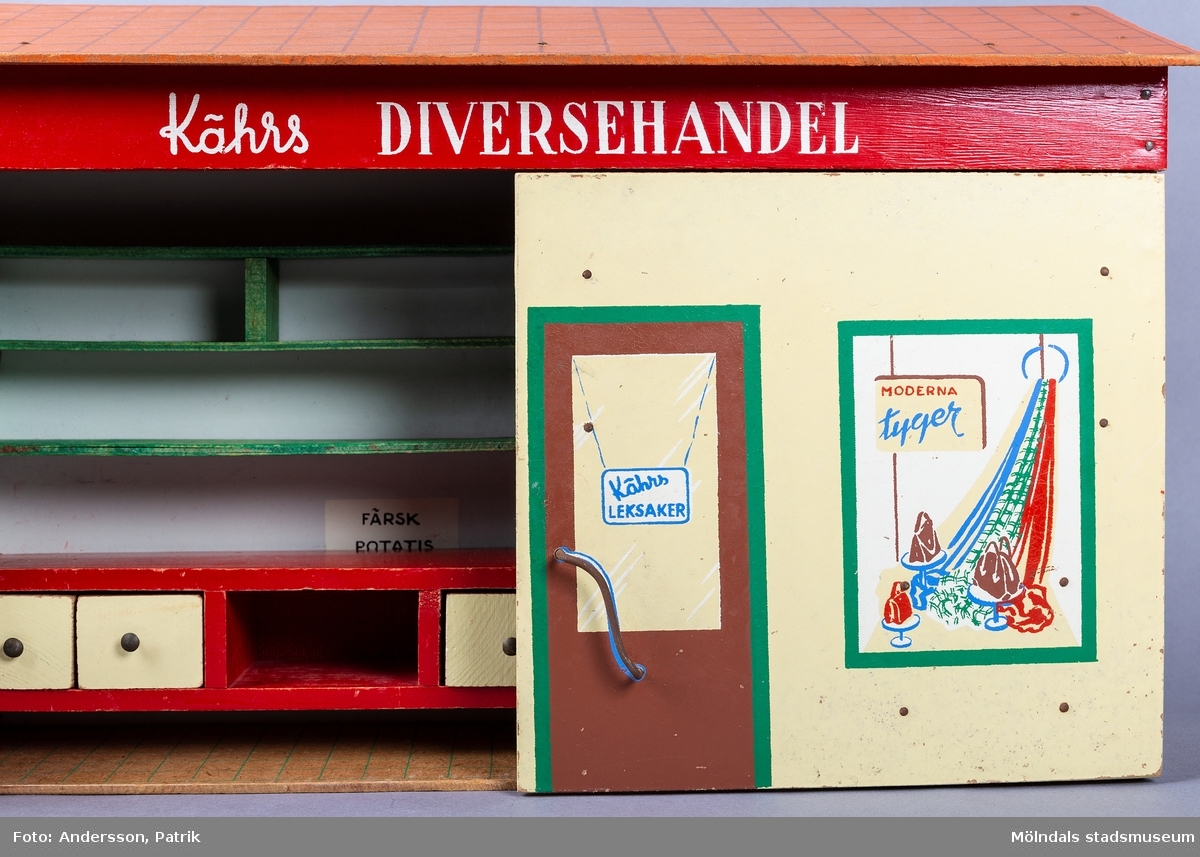 Leksak i trä i form av en liten affär. På framsidan finns texten "Kährs Diversehandel", dekor i form av två skyltfönster och en liten dörr med ett handtag i metall. Fronten består av två luckor går att öppna. Inuti finns en butiksinredning med gröna hyllor och en lös röd butiksdisk med sex gula utdragbara lådor.
Allt är tillverkat i trä. Affärens utsida är målad i en ljusgul färg, med ett tak som är bemålat med tegelröda tegelpannor. Under takåsen löper en röd list med texten "Kährs Diversehandel" målat med vita bokstäver.
Leksaksbutiken är tillverkad av Kährs. Företaget grundades 1857 och tillverkade till en början leksaker och möbler, men tillverkningen övergick efter andra världskriget alltmer till att omfatta endast dörrar och golv.