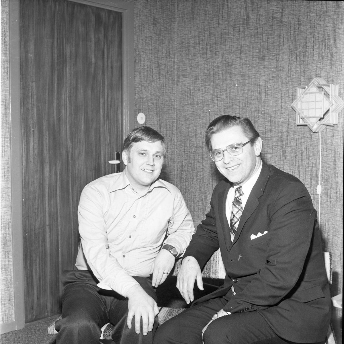 Två glada män sitter intill varandra i ett rum med strukturtapet och fanerad dörr i samma ton. Mannen till höger bär kostym.