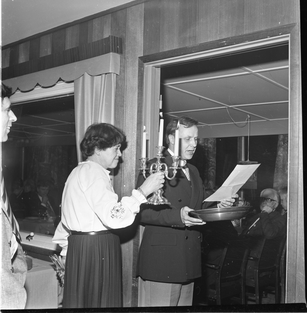 Karin Wenner håller upp en tänd trearmad ljusstake för Allan Svensson som håller ett tennfat (troligen) och läser från ett papper. Till vänster skymtar Tom Sonesson.