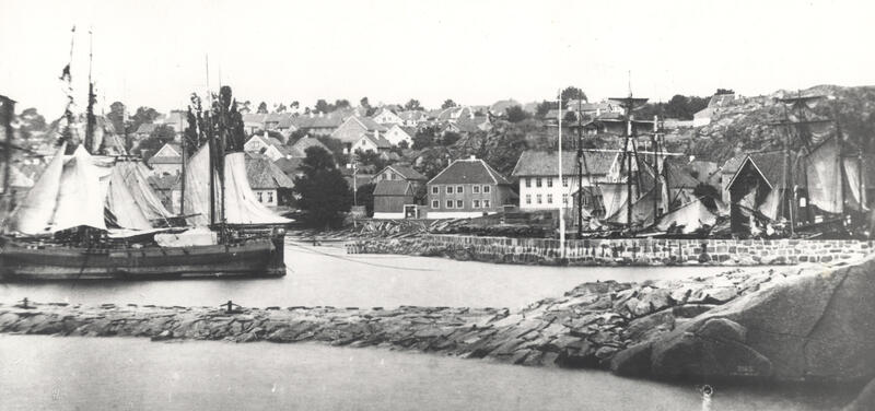 Sort-hvitt-bildet viser en mursatt molo med seilskuter. I bakgrunnen ser vi trebygninger (Foto/Photo)