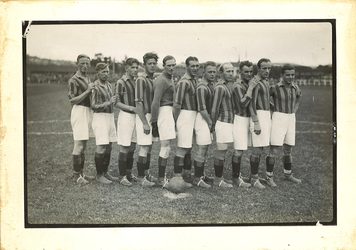 Sort-hvit-bildet viser 11 unge menn som står åp rekke. De har hvite, litt lange shortser, stripete trøye med v-hals og krage og like ensfargede sko og strømper på bena. Ballen er mørk.