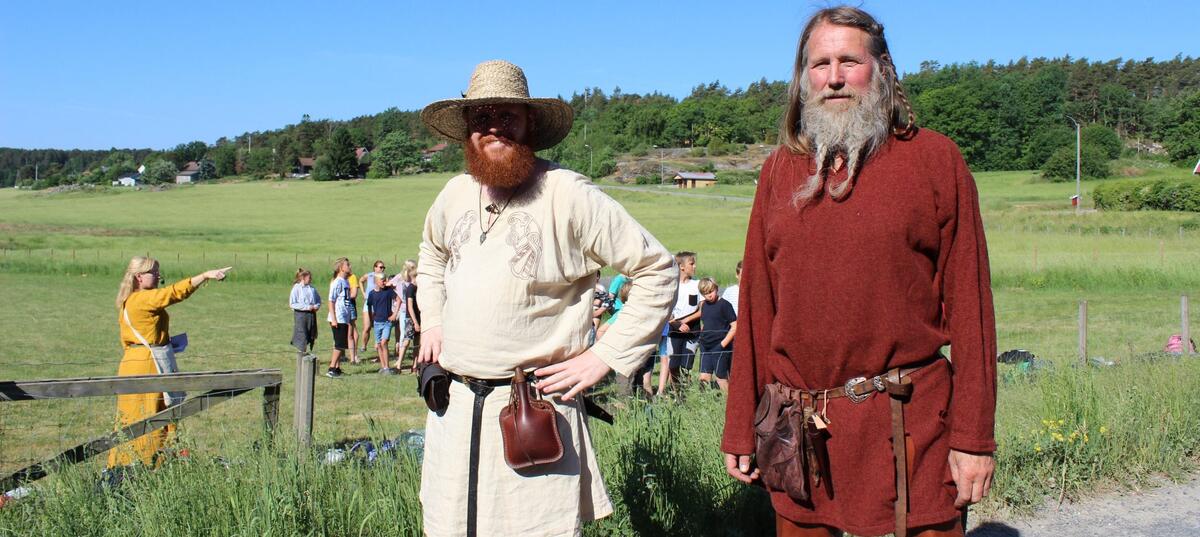 Bildet viser to vikingkledde menn med skjegg. I bakgrunnen er det grønne sletter, blå himmel og noen mennesker i aktivitet (Foto/Photo)