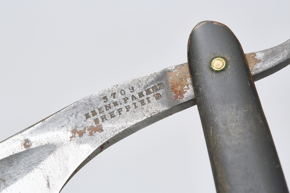 Barberskrinet (a) er umalt, skyvelokk rikt utskåret på sider og lokk. Barberkniv (b) med hornskaft og stålblad, bladet kan foldes inn i skaftet. Reserveblad (c).