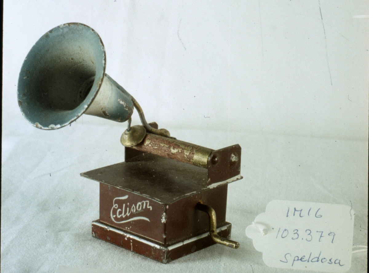 Speldosa i form av trattgrammofon, av metall, fyrkantig låda med vals av trä samt stor tratt i grönt, vev av metall på ena sidan. Lådan målad i brunt. "Edison" målat i vitt på framsidan. Fabrikstillverkad.