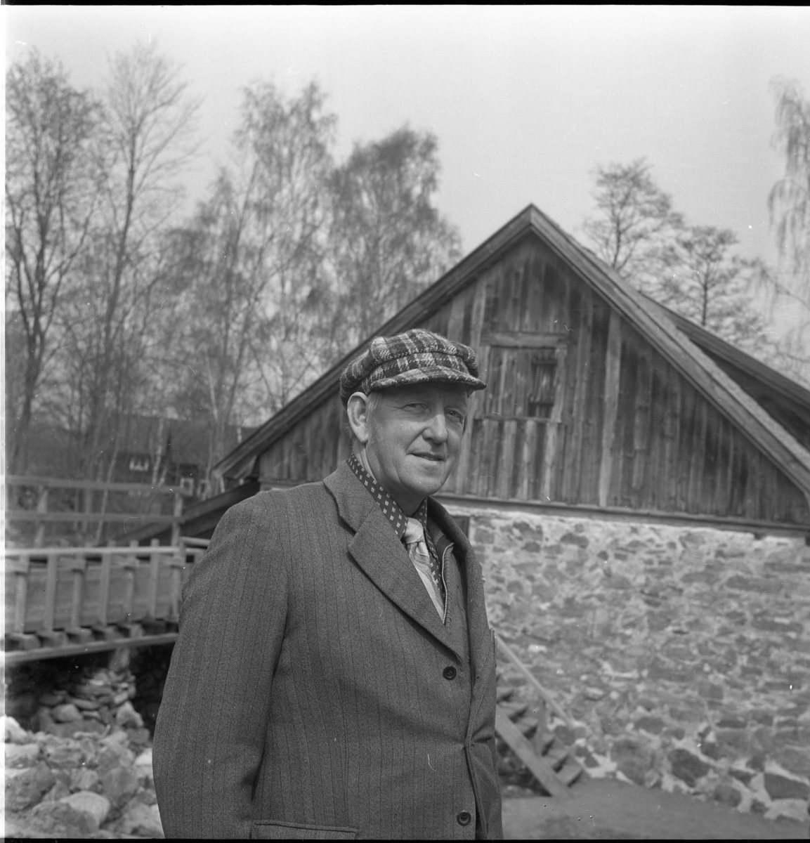 Porträtt av en man med keps, slips och blazer stående vid Rasmus kvarn östra gavel.