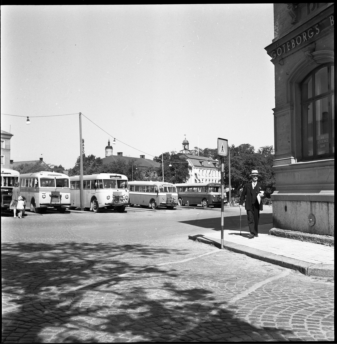 Arvid Åman promenerar vid Lilla torget. Augusti 1950. I bakgrunden syns parkerade bussar på torget. Kaboms skorsten, Stadskyrkans torn samt slangtornet vid Väktaregården sticker upp över hustaken.