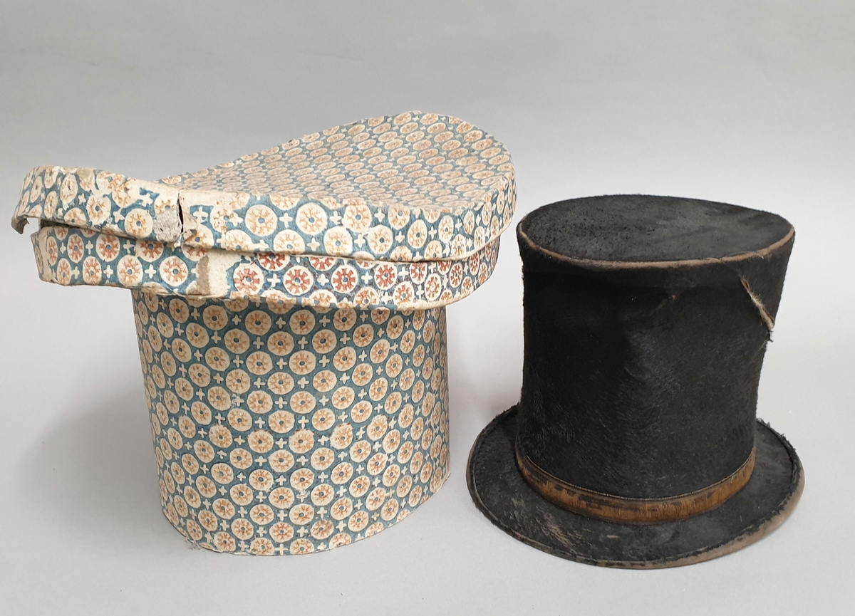 Hatt med flosset overflate og silkebånd rundt pullen. Til hatten følger en hatteeske av kartong som er formet som hatten, og trukket med mønstret papir.