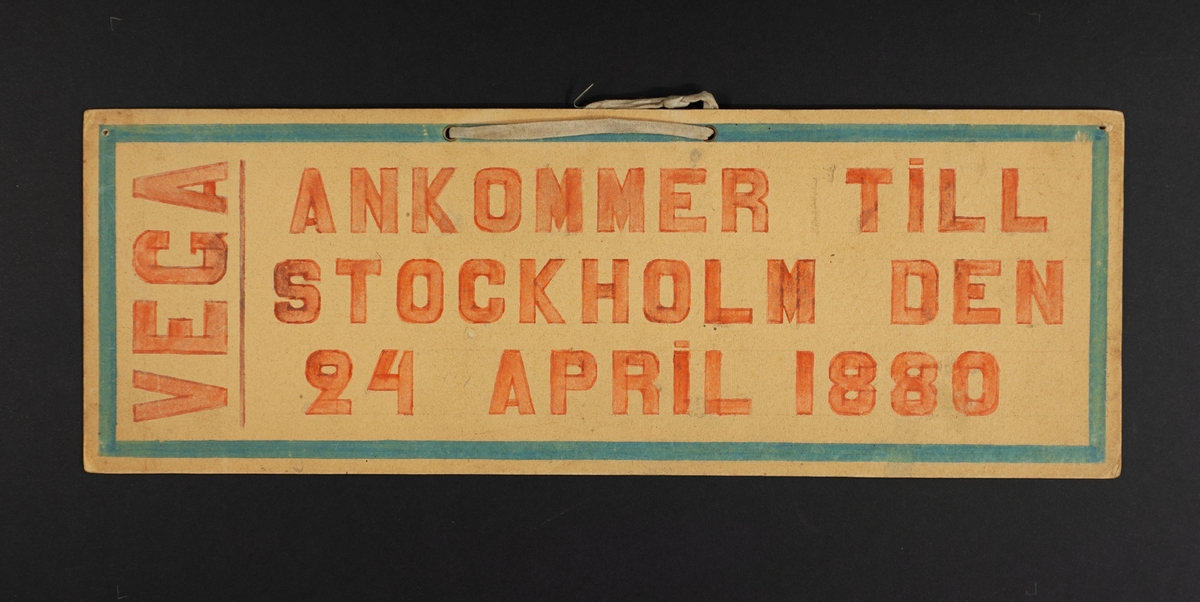 Handtextad skylt i rött och blått av papp, med textilt upphängningsband: "Vega ankommer till Stockholm den 24 April 1880". På baksidan påskrift "Enke Kejsarinan af Tyskland".