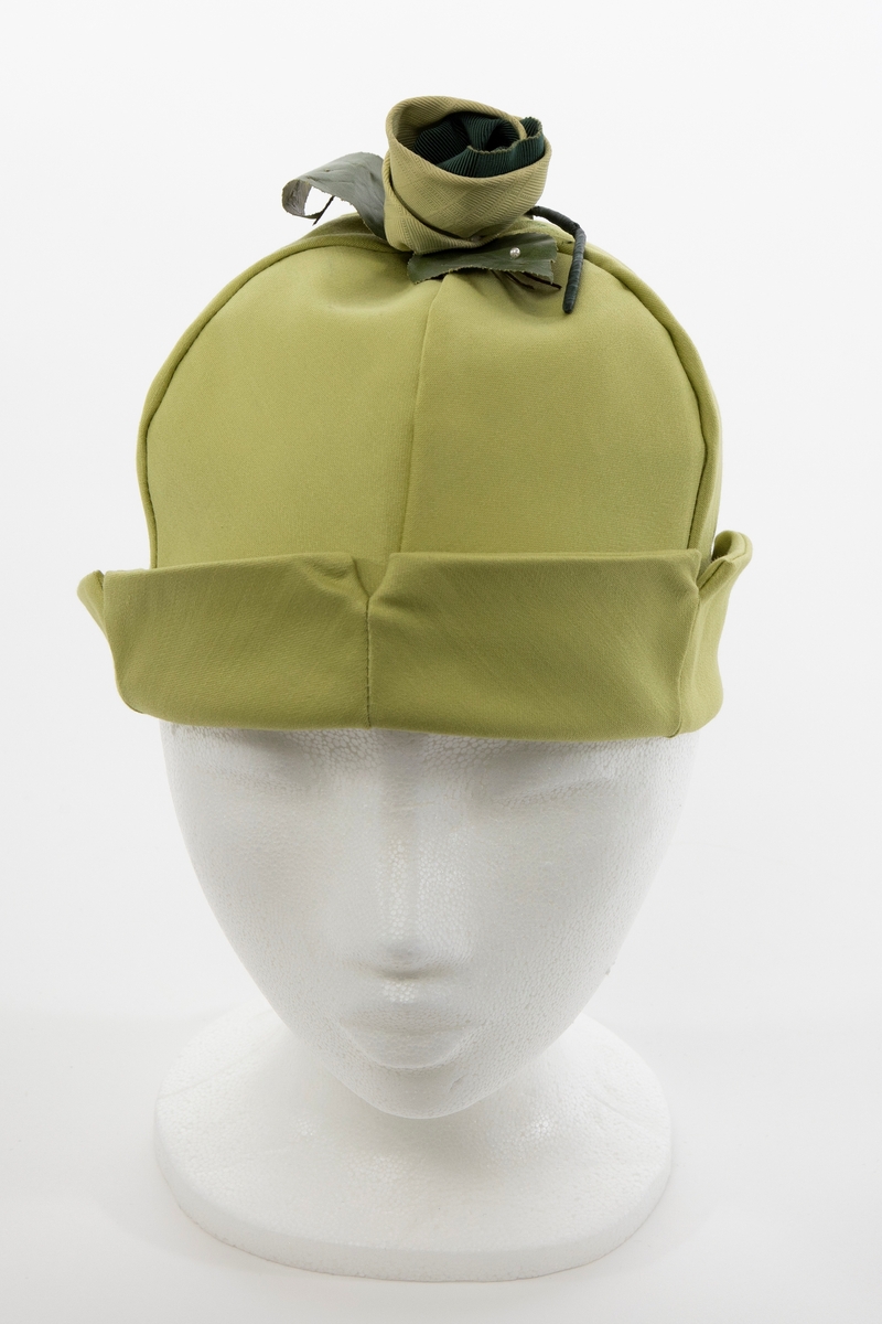 Klokkeformet, sommerlig hatt med opprett nederst. Satengvevd, halvblankt stoff. Hatten er dobbel, dvs at den er sammensydd av 2 x 6 sektorer, slik at den har lik konstruksjon innvendig som utvendig. Hatten skal være oppbrettet, og det er korte splitter i nederkant mellom hver sektor. Oppbrettet gir det en takket kant på oppbretten. På toppen av hatten er det påsydd en tekstilrosett, en slags "Dior-rose", konstruert av et limefarget syntetstoff med stripet og rutet overflatestruktur, omkring et innvendig, mørk grønt, ripsartet silkebånd. Mellom rosen og hatten er det festet 3 mørkegrønne blader av sterkt appretert tekstil, med stilker av wirer og én tykkere, vokset hovedstilk. Det erne bladet er prydet med en påsydd, ørliten. perlemorskimrende perle. Det kan se ut som også de 2 andre bladene har hatt en liten perle.