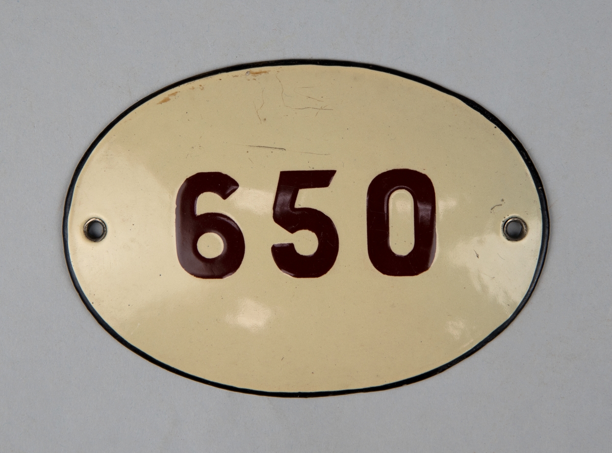 Oval vagnsnummerskylt av emaljerad metall. Vit botten med "650" i brunt. Svart kant runt kanten. Två hål för fastsättning.