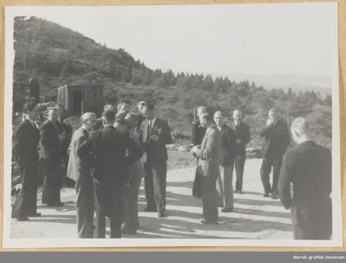 Utendørsmotiv. En gruppe menn i dress står på en grusvei eller -slette. Spredt skogvekst og en hytte eller uthus i bakgrunnen. Fotografert i forbindelse med Vestlandsk Trykkerstevne i Bergen, 1946.