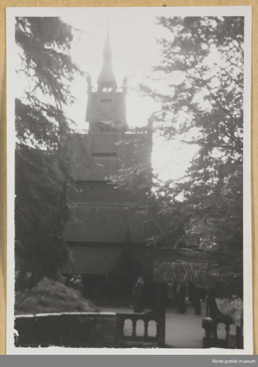 Bygning. Fantoft stavkirke, sett fra utsiden av porten med trær på begge sider. Noen mennesker skimtes foran kirken. Fotografert i forbindelse med Vestlandsk Trykkerstevne i Bergen, 1946.