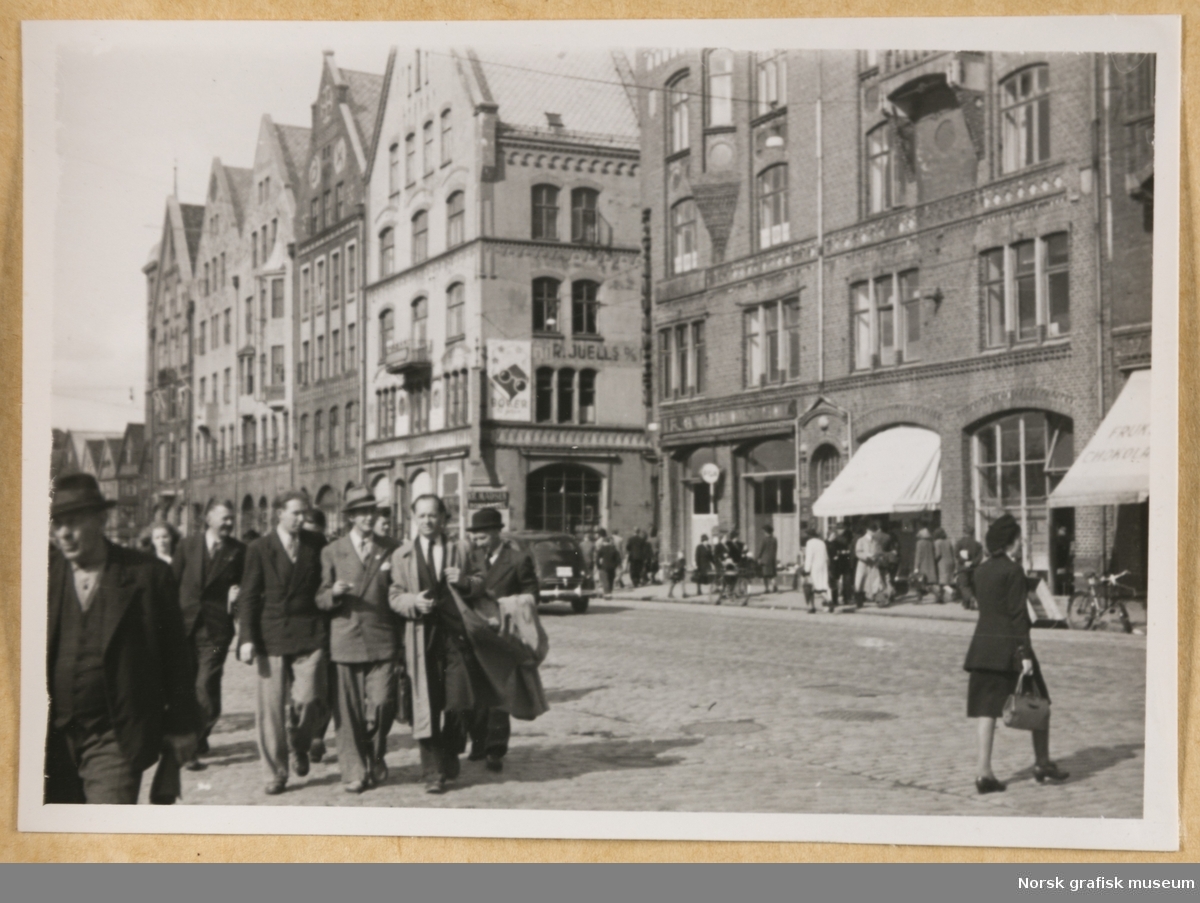 Gatemotiv. Murbygninger i fem-seks etasjer med brosteinsbelagt gate eller plass foran. I forgrunnen en gruppe menn i dress. På en av bygningene skimtes firmanavnet R. Juell. Fotografert i forbindelse med Vestlandsk Trykkerstevne i Bergen, 1946. 