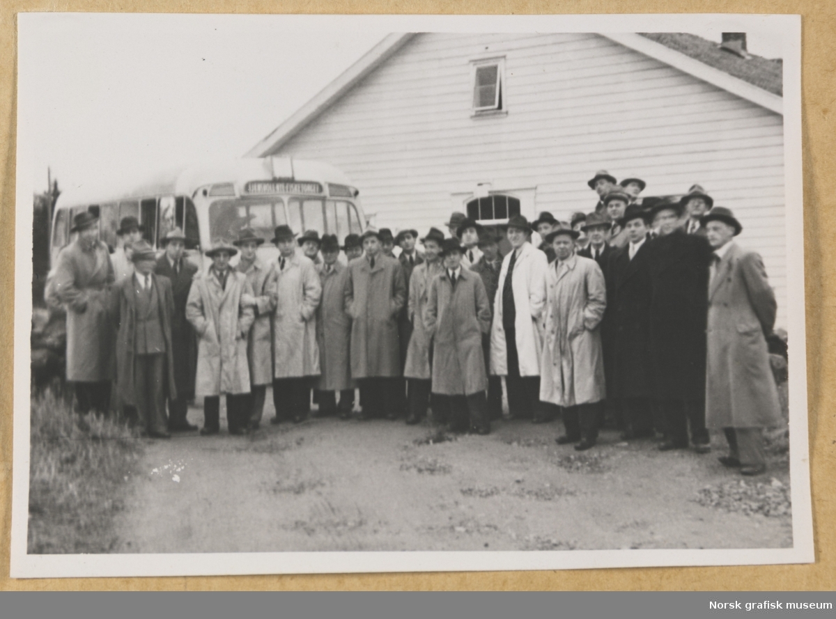 Utendørs gruppebilde. Rundt tretti menn i frakk og hatt står oppstilt foran en buss og en trebygning. Fotografert i forbindelse med Vestlandsk Trykkerstevne i Stavanger, 1948.