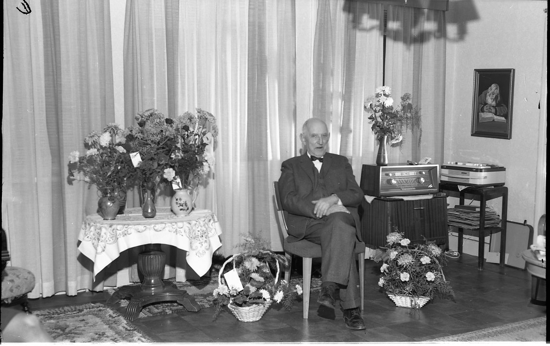En äldre man sitter i en stol. På ett bord intill och på golvet står blomsterbuketter. Mannen heter Möller och fyller 75 år. Till höger om mannen står en radio på ett litet skåp och därintill en rullbandspelare på ett bord.