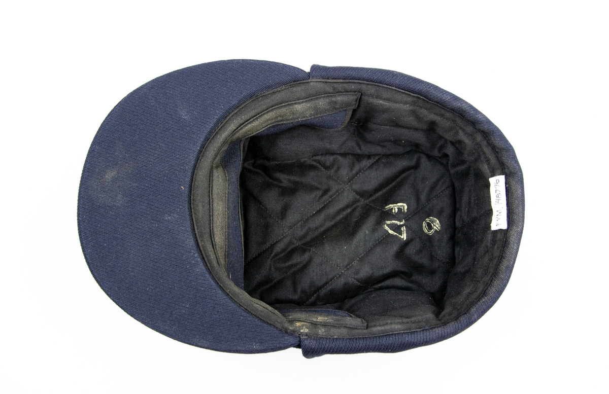 Vintermössa, blå m/1954 med skärm och öronskydd.  Över skärmen sitter mössmärke m/51. Fodrad med mörkblått tyg,