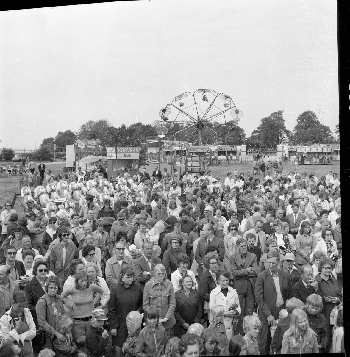 En folksamling står och tittar upp mot en scen, i bakgrunden karuseller, lottstånd och korvbar.