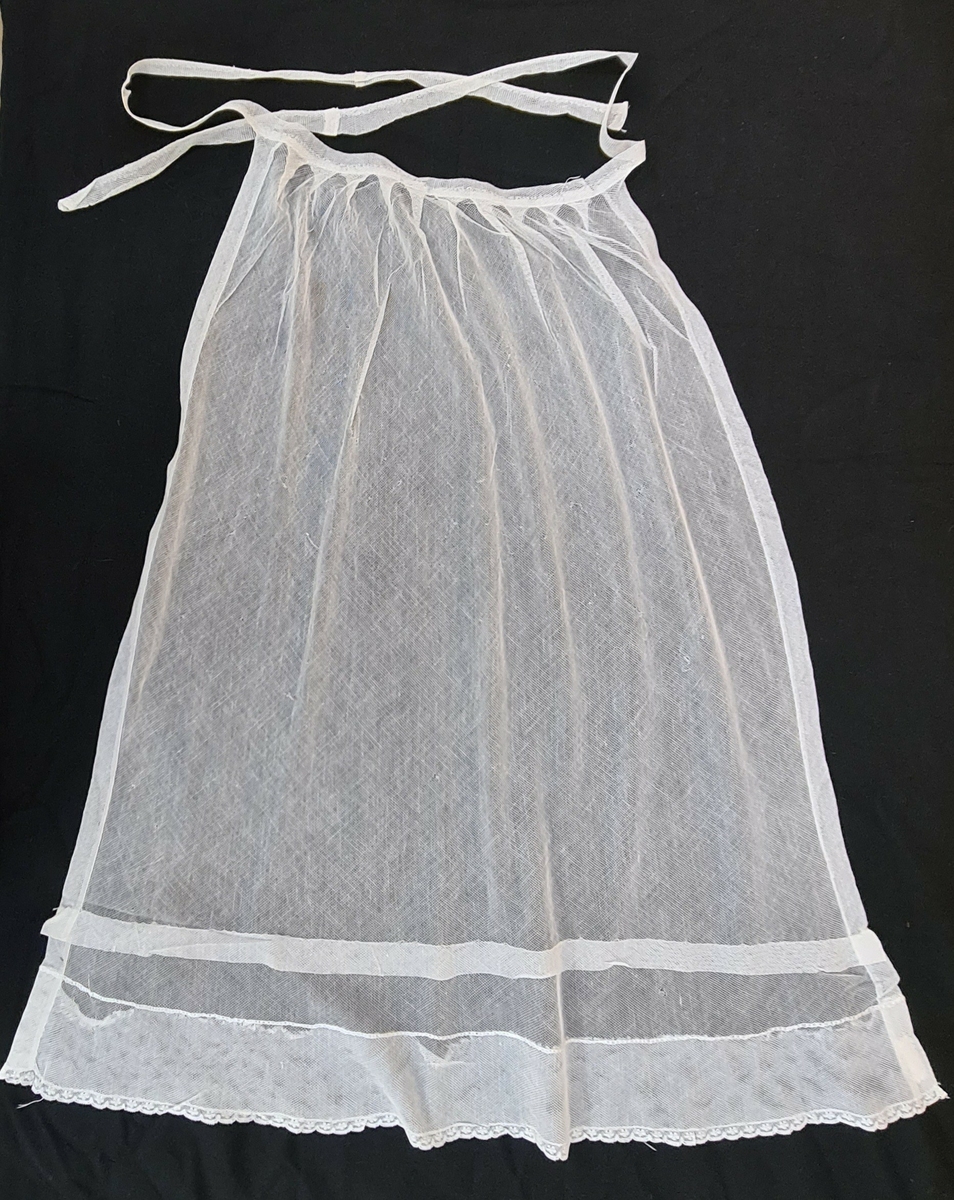 Vitt midjeförkläde i tyll med spets nedtill. Buret före 1915 av Berta Elow (mor till givarna).