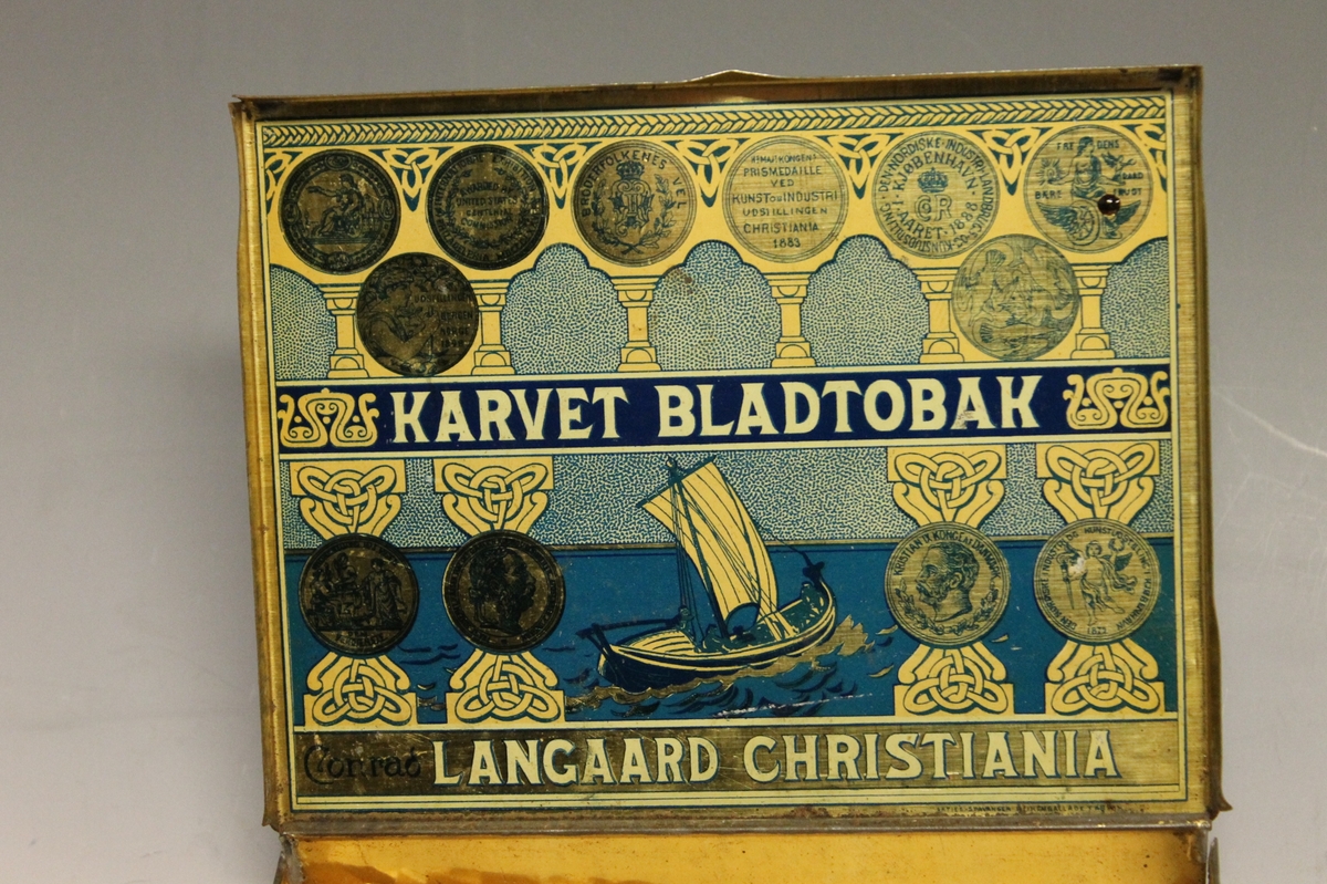 På lokket og på innsiden av lokket er det et trykkbilde av en nordlandsbåt med råseil. Båten, sjøen og himmelen er omkranset av "stolper" og arkader" med medaljer/segl for utmerkelser. På midten et midtparti hvor det står "Karvet Bladtobak". Nederst står det "Conrad Langaard Christiania". Blanding av keltisk og vikingeornamentikk.