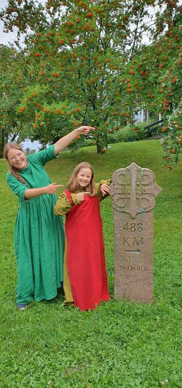 To unge jenter i middelalderkjoler står og peker på en pilegrimsmilestein med påskriften "488 km til Nidaros"