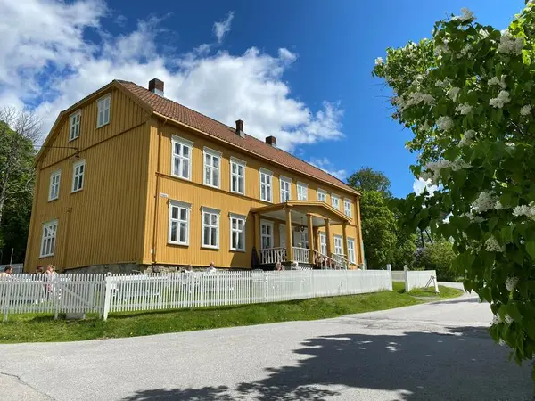 Aamodtgården i sola. Gult, staselig bygg med to etasjer og et loft. Huset og hagen er inngjerdet av et hvitt stakittgjerde. Til venstre i bildet, i forgrunnen, er det en syrin med hvite blomster. Fotografiet er tatt i fint sommervær.