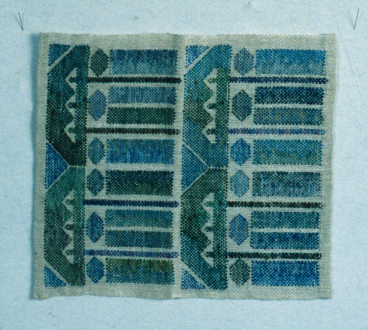 Duk i HV-teknik, vit och oblekt botten med mönster i gröna och blå nyanser, brunt och svart. Handfållad i två sidor.