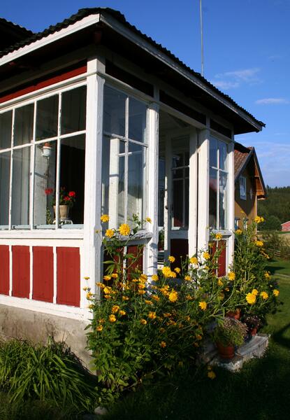 Glassverandaen til Vestun på Almenninga. Rødt bygg med hvitkarmede vinduer. Gule blomster ved inngangstrappa til glassverandaen.