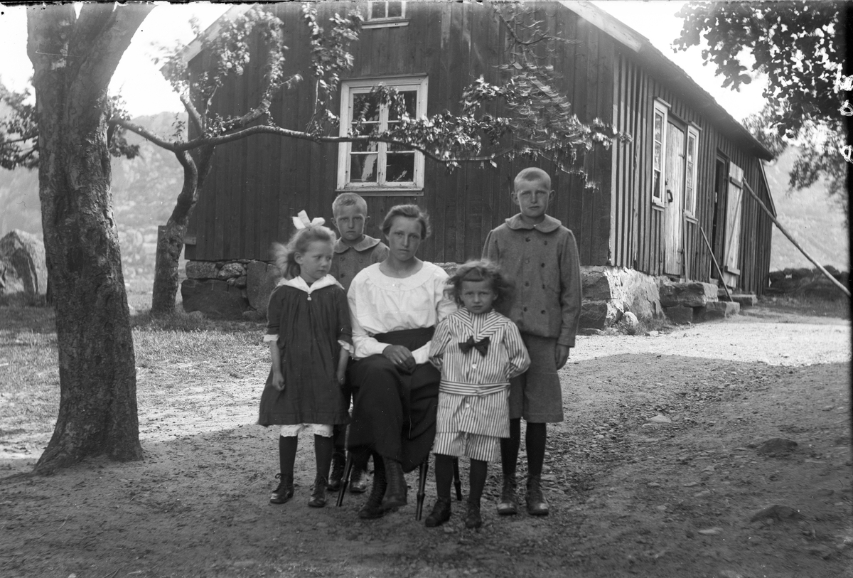 En mor, eller kanske en storasyster, sitter omgiven av fyra barn i trädgården vid ett gårdshus. De två äldsta pojkarna är klädda likadant, medan den minste är klädd i kortbyxor och kolt med sjömanskrage, allt i samma randiga tyg. Flickan har en mörk klänning med vita spetsar på krage, ärmar och nedtill. 
(Se även bild nr GEA134)