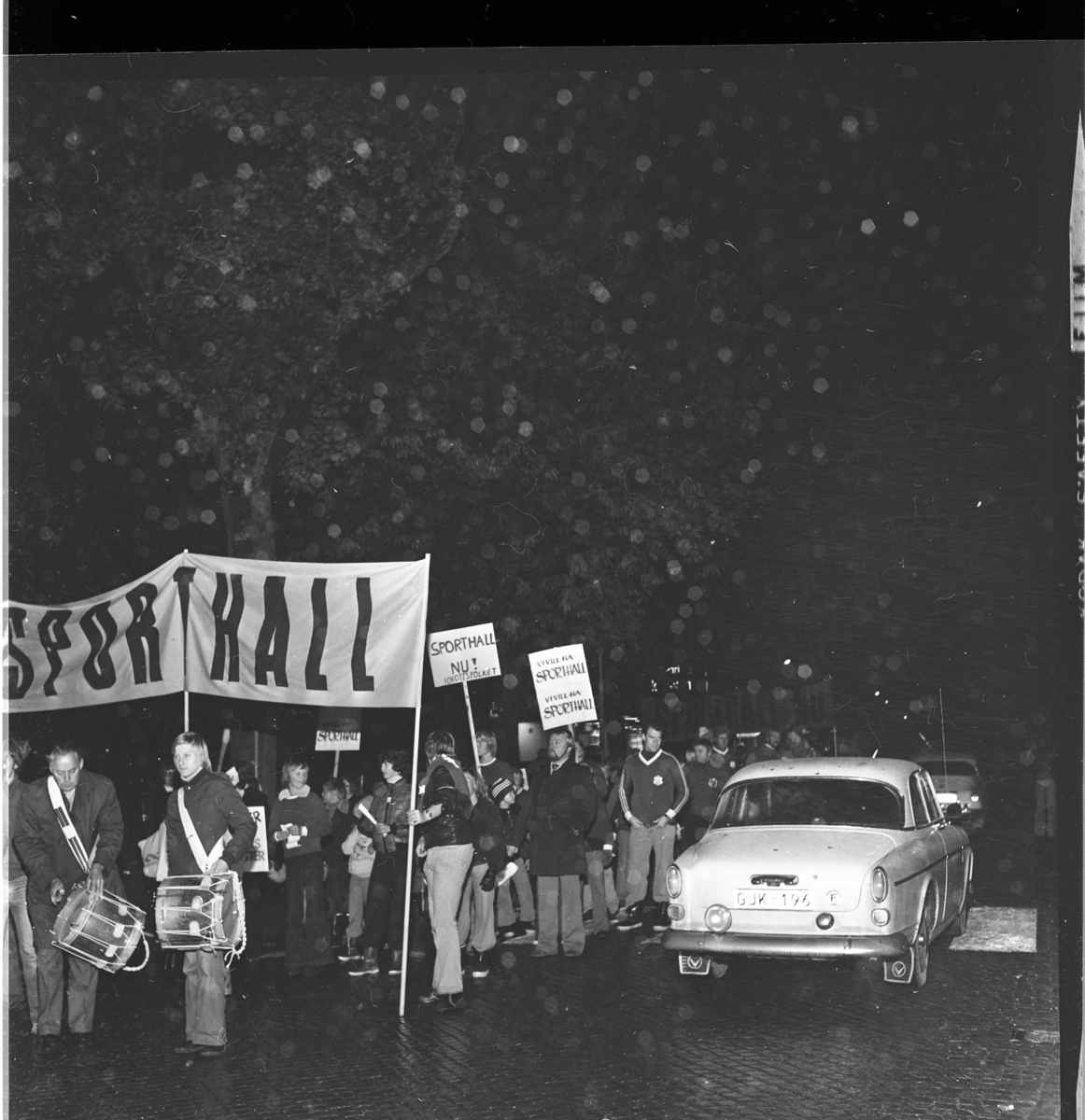 Ungdomar och vuxna med banderoll och plakat om "Sporthall". Trumslagare i täten. Grännabor demonstrerar för att få en sporthall till Gränna. Det är oktober, mörkt och blött på vägen. En Volvo Amazon passer demonstrationståget.