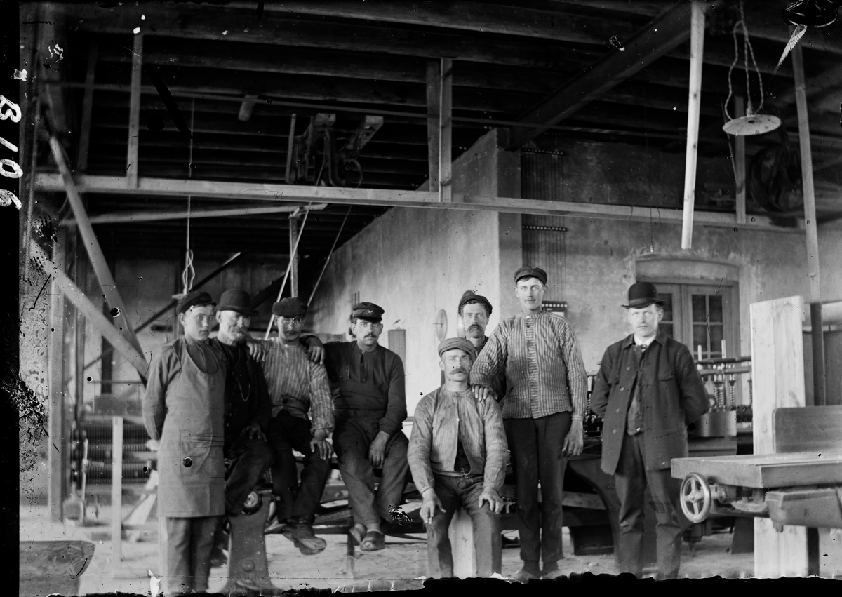 Interiör av fabrik med fabriksarbetare i olika åldrar. I bakgrunden syns remdrift till någon maskin. Två av männen bär kubb, övriga skärmmössor och klädseln varierar.