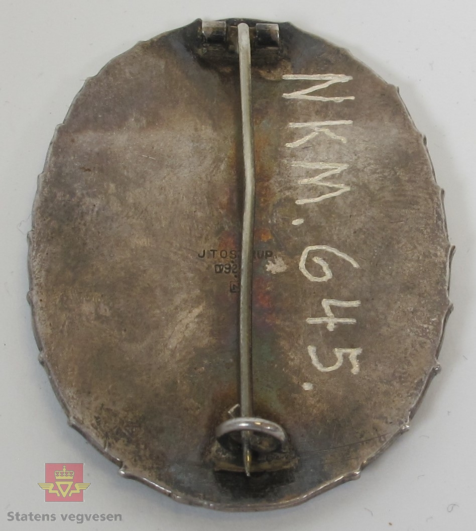 Jakkemerke laget av sølv formet som en oval med nål på baksiden for festing.