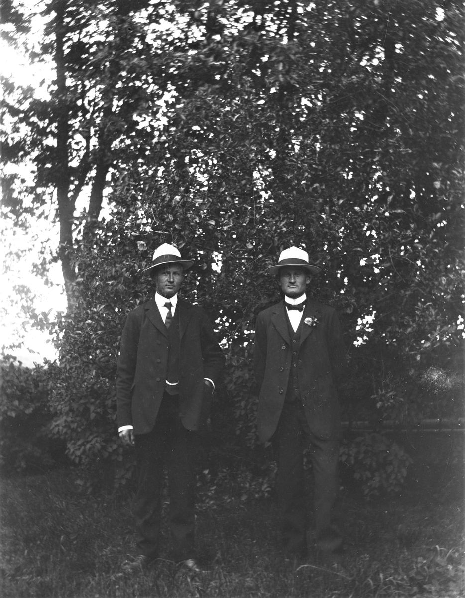 Två yngre män står finklädda utomhus med likadana ljusa, toppiga hattar med mörka band och den ene bär en blomma på kavajslaget.