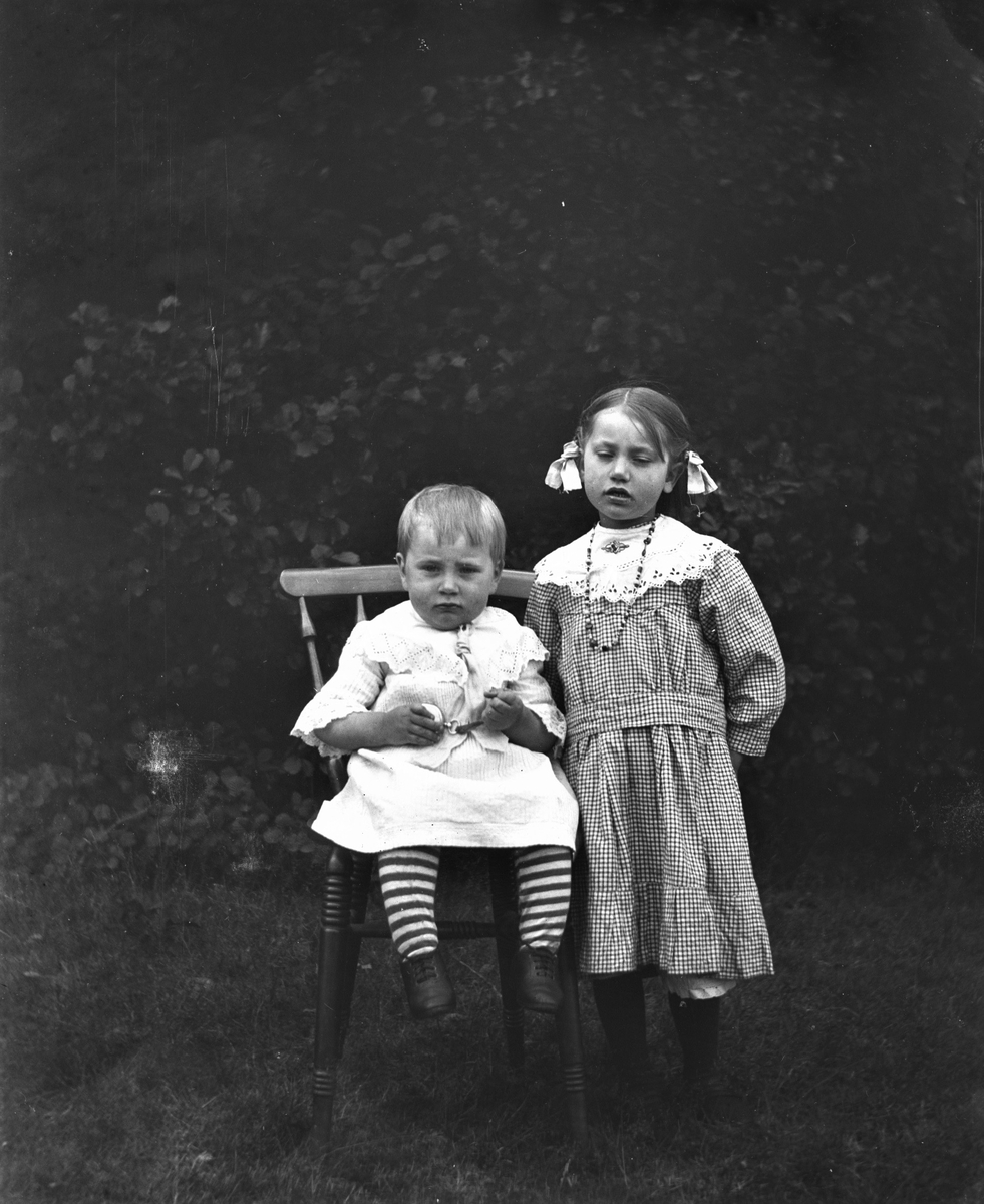 Två små barn fotograferade utomhus. Den lille sitter på en pinnstol och håller i ett fickur, den äldre flickan står bredvid. Hon har en rutig klänning med spretskrage, pärlhalsband, brosch och en frisyr med rosetter vid öronen. (Se bild på övriga familjen GEB201)