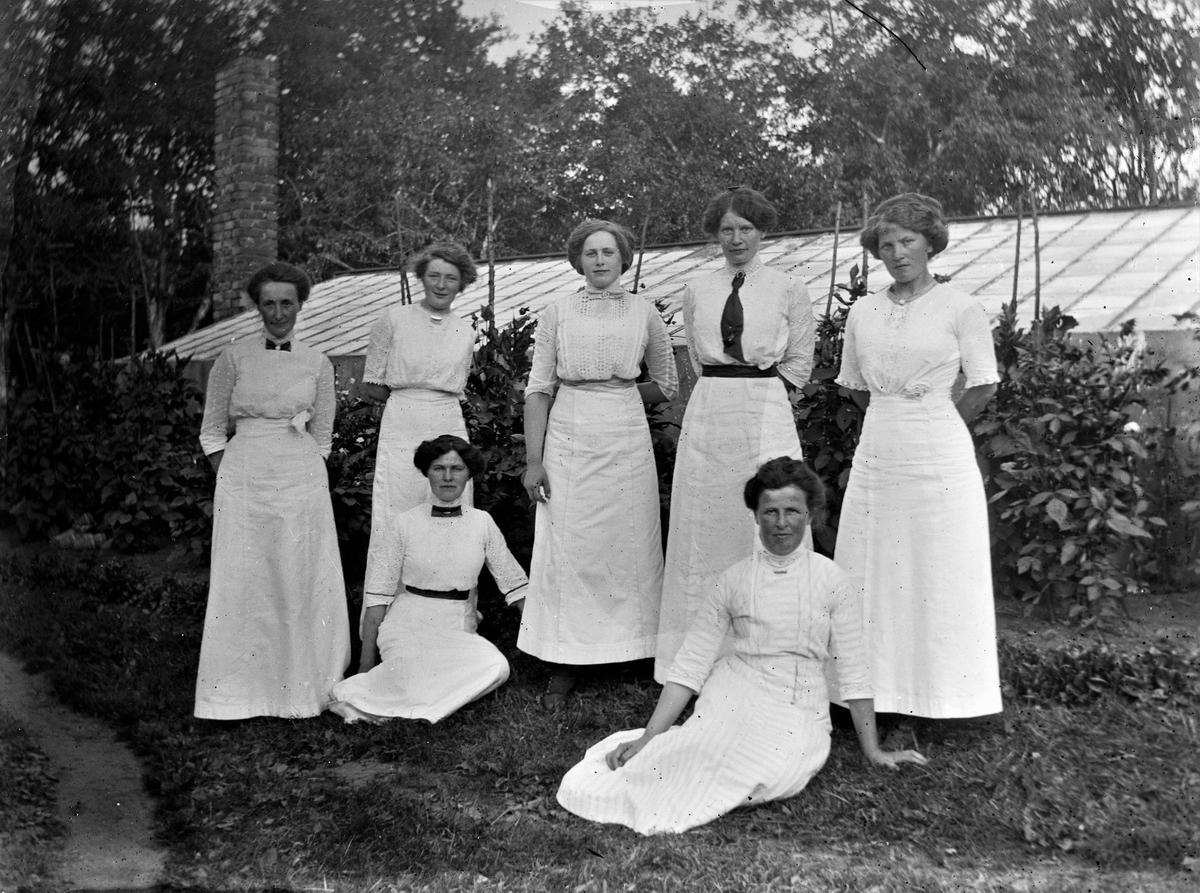Sju vitklädda kvinnor vid en rabatt med prunkande dahlior och bakom dem ett växthus med skorsten i bortre änden.