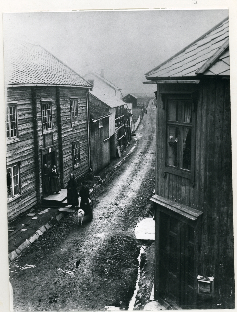 Til venstre i bildet sees Fattighuset på Røros med fire av beboerne stående på trappa. Midt i bildet går "Storgjellen"/Peder Hiort gata vestover. Sett fra Nilsen-hjørnet